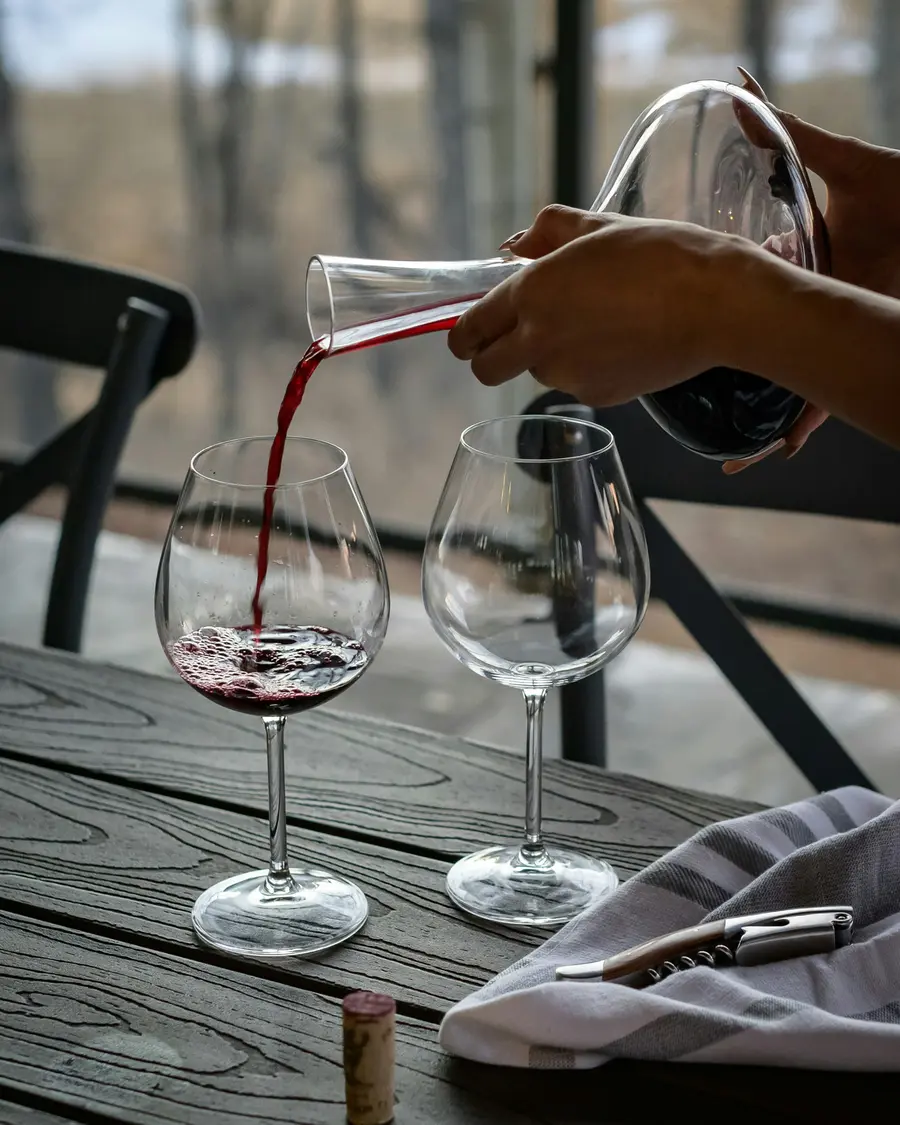 Red wine being poured from a decanter into a Burgundy glass on a wooden table.